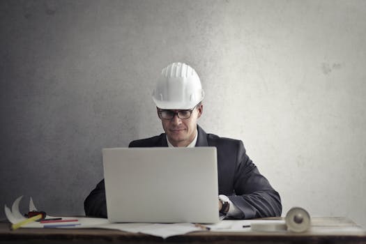 Professional engineer wearing hardhat working on construction plans at desk with laptop in office.